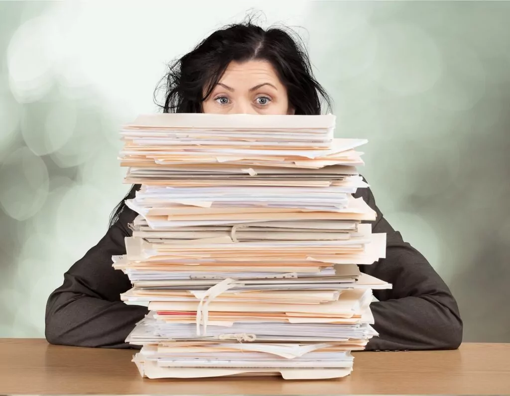 A woman is sitting at a desk and peering over a large stack of documents and folders.